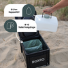 Schwarzer Kasten mit offenem Deckel und sichtbarer grüner Innentasche, Markenaufdruck 'BOXIO'; Hand hält weißen Kanister mit grüner Verschlusskappe über der Öffnung; sandiger Strand mit Dünengras im Hintergrund; zwei weiße Info-Etiketten mit den Text