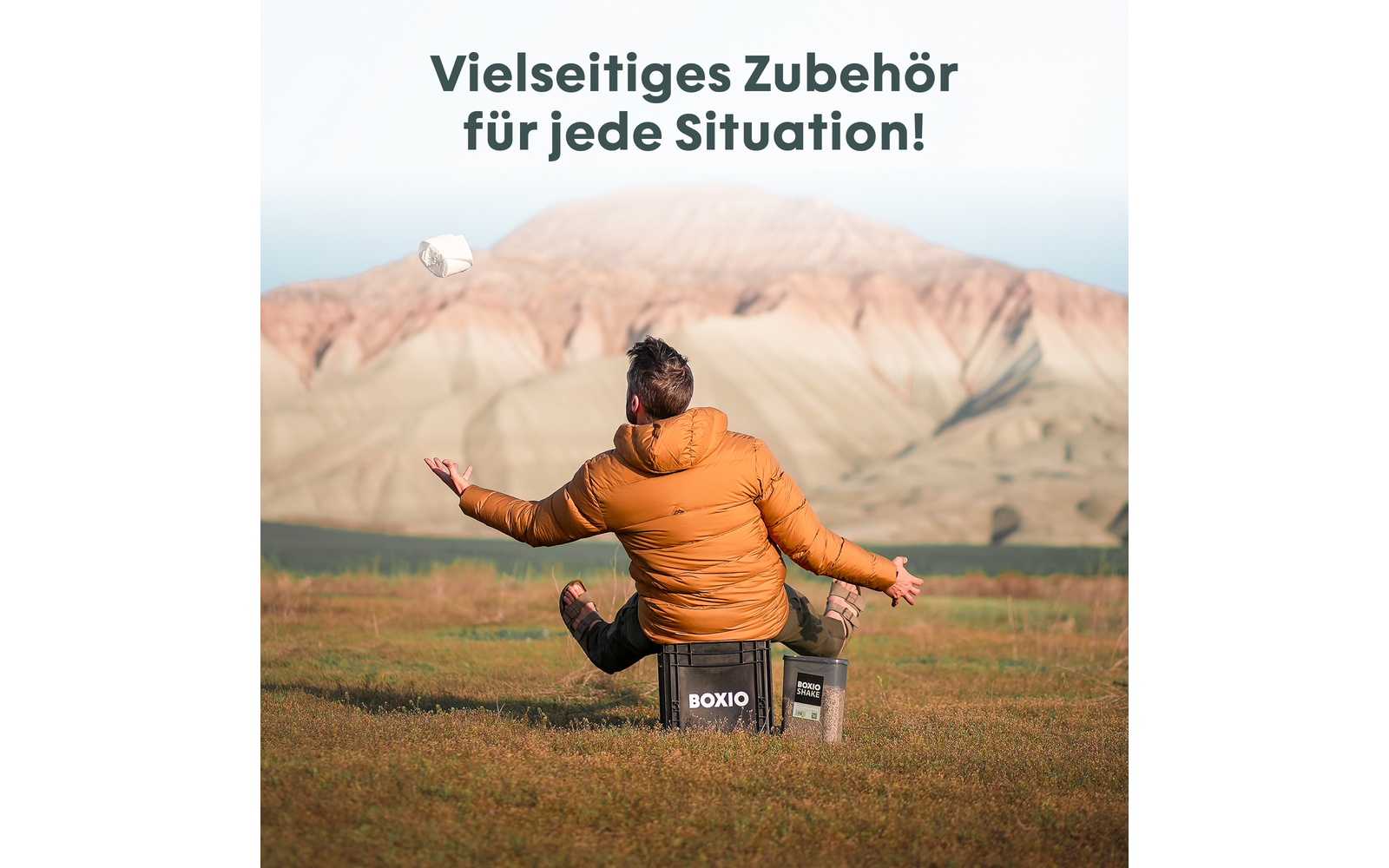 Person in orangener Steppjacke sitzt mit dem Rücken zur Kamera auf einer schwarzen Box mit der Aufschrift "BOXIO" auf einer Wiese und wirft ein weißes Objekt; Berge im Hintergrund