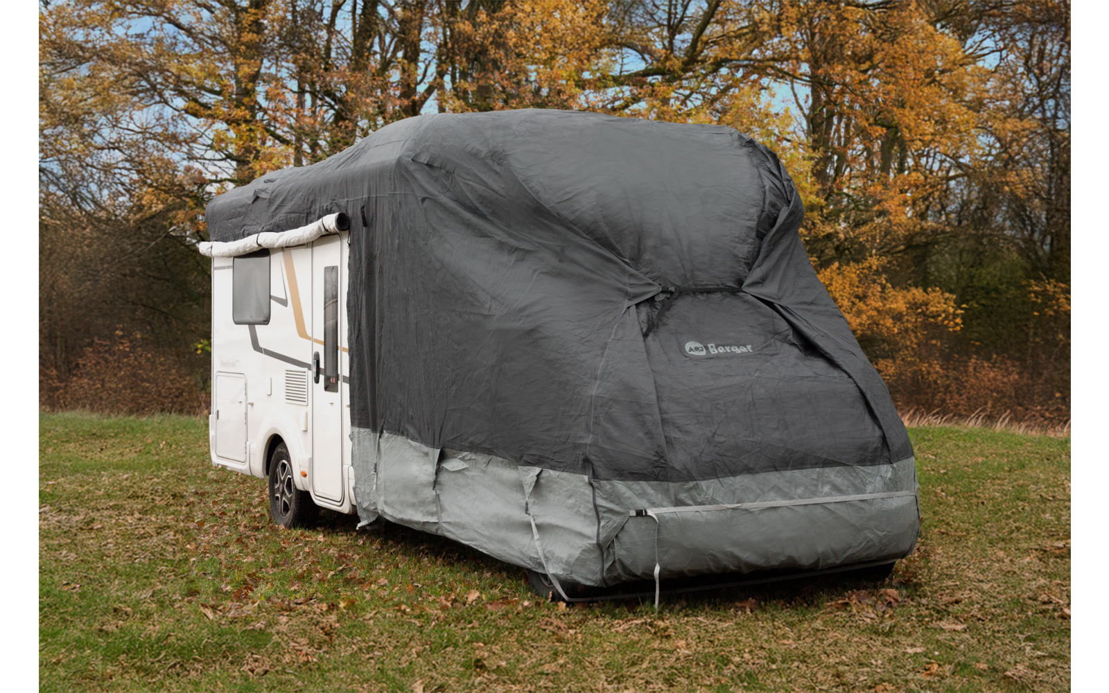 White motorhome covered with a gray protective cover on grass, trees with yellow-brown leaves in the background
