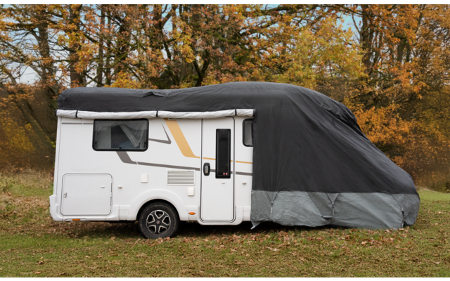 White motorhome on grass, front half covered with grey and black protective cover, window, door and wheel visible, trees with autumn foliage in the background