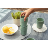 Mano recoge una taza verde apilada sobre una mesa de madera gris; cuenco con limones, pequeña planta en maceta y otra taza verde sobre un posavasos blanco visibles