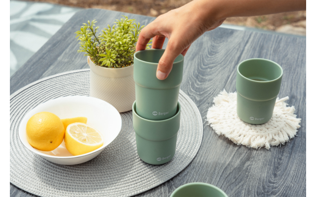 Mano recoge una taza verde apilada sobre una mesa de madera gris; cuenco con limones, pequeña planta en maceta y otra taza verde sobre un posavasos blanco visibles