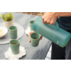 Mano y antebrazo de una persona vertiendo líquido desde una jarra verde en una taza verde sobre una mesa de madera oscura; dos tazas verdes adicionales, posavasos blancos de ganchillo, una pequeña planta en maceta blanca y un cuenco con rodajas de limón visibles