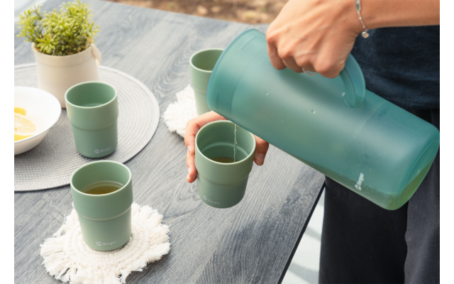 Mano y antebrazo de una persona vertiendo líquido desde una jarra verde en una taza verde sobre una mesa de madera oscura; dos tazas verdes adicionales, posavasos blancos de ganchillo, una pequeña planta en maceta blanca y un cuenco con rodajas de limón visibles