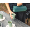 Manos vertiendo líquido marrón desde un termo verde en una taza verde sobre una mesa de madera gris; otras tazas verdes, posavasos blancos y una silla negra en el fondo