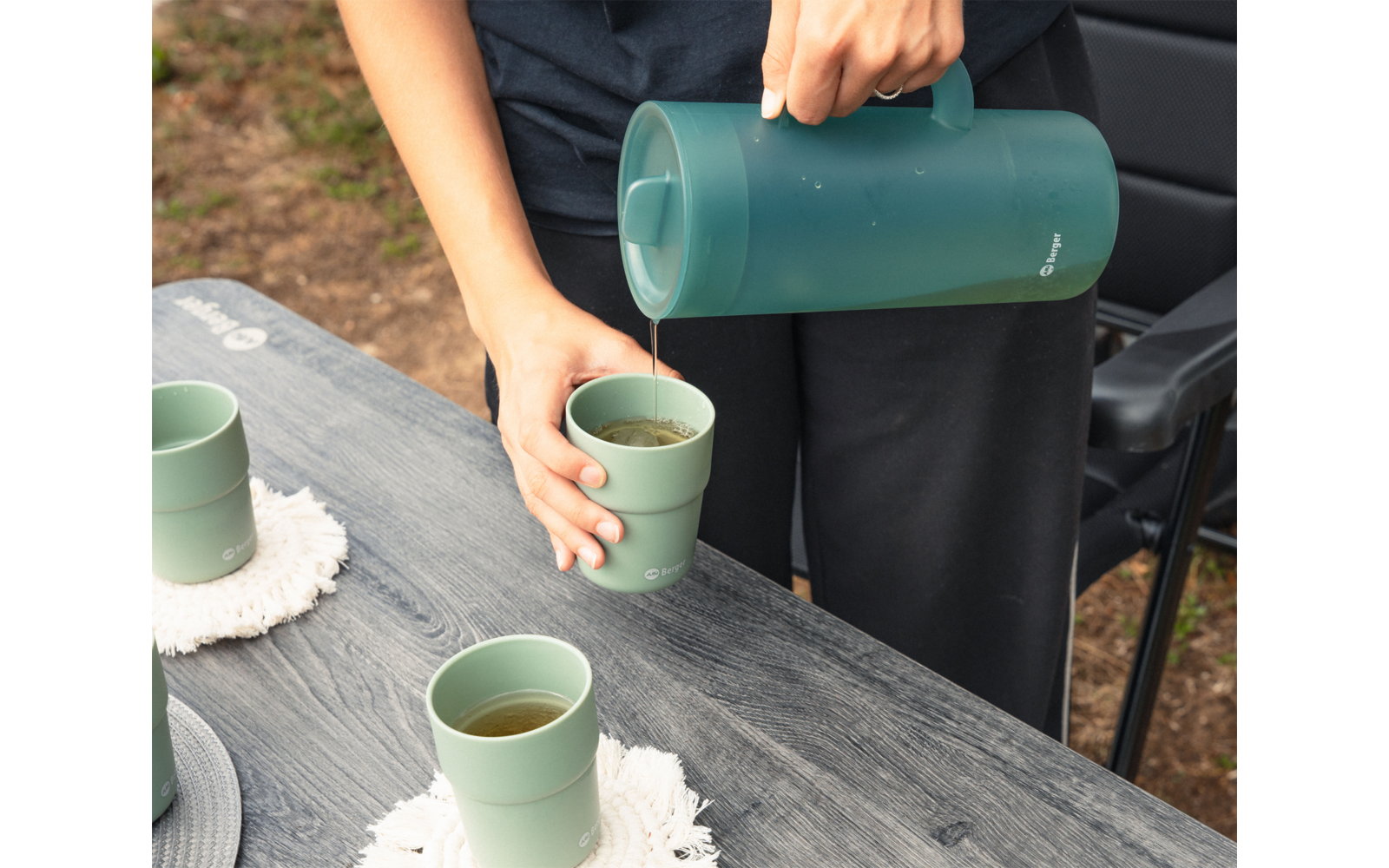 Manos vertiendo líquido marrón desde un termo verde en una taza verde sobre una mesa de madera gris; otras tazas verdes, posavasos blancos y una silla negra en el fondo