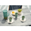 Superficie de mesa de madera gris con a la izquierda una jarra turquesa con tapa, tres tazas turquesa (dos delanteras sobre posavasos blancos, una sobre un individual redondo gris claro), sobre el individual un cuenco blanco con mitades de limón y una maceta blanca pequeña con una planta verde, respaldo de silla azul oscuro visible en la parte superior