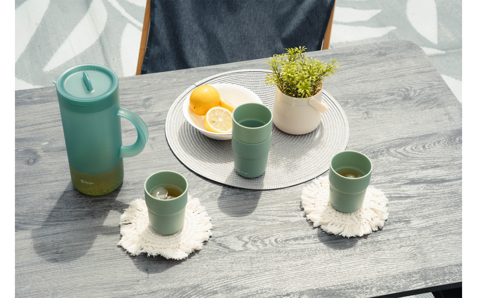 Superficie de mesa de madera gris con a la izquierda una jarra turquesa con tapa, tres tazas turquesa (dos delanteras sobre posavasos blancos, una sobre un individual redondo gris claro), sobre el individual un cuenco blanco con mitades de limón y una maceta blanca pequeña con una planta verde, respaldo de silla azul oscuro visible en la parte superior