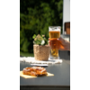 Twee glazen bier, een hand houdt een glas vast, pretzels liggen op een witte tafel, groene planten op de achtergrond