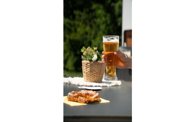Twee glazen bier, een hand houdt een glas vast, pretzels liggen op een witte tafel, groene planten op de achtergrond