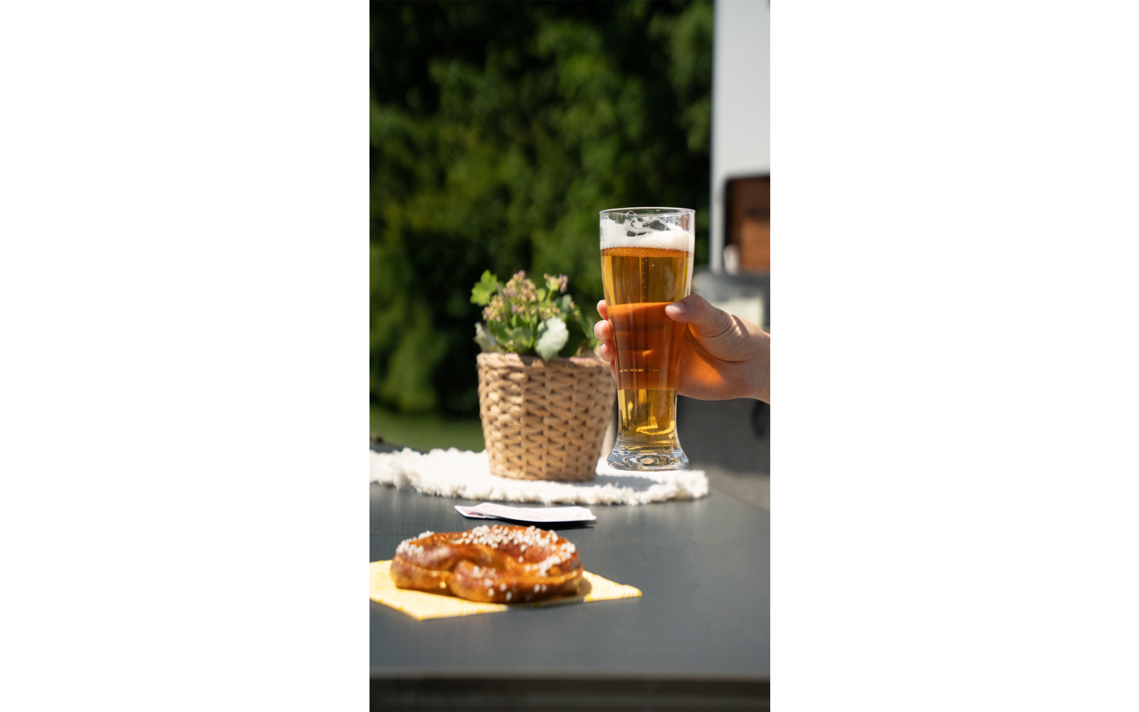 Twee glazen bier, een hand houdt een glas vast, pretzels liggen op een witte tafel, groene planten op de achtergrond