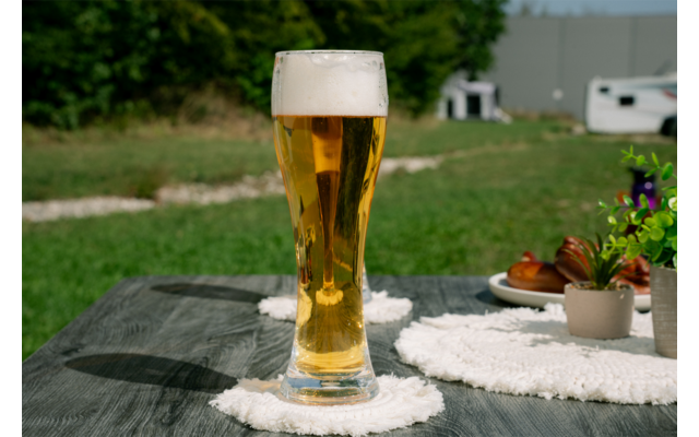 Grand verre de bière claire avec mousse sur une table à l'extérieur, sous-verre blanc, pelouse et caravane en arrière-plan