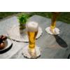 Deux verres hauts contenant un liquide doré et de la mousse sur des dessous de verre ronds blancs à franges sur une table en bois foncé ; petit pot avec plante, assiette avec bretzels au bord de la table, pelouse en arrière-plan