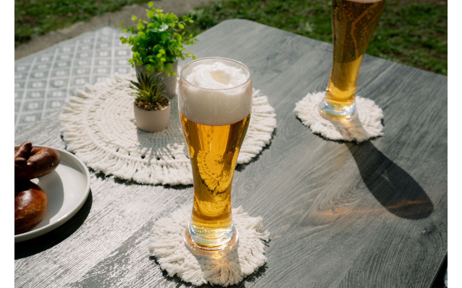 Deux verres hauts contenant un liquide doré et de la mousse sur des dessous de verre ronds blancs à franges sur une table en bois foncé ; petit pot avec plante, assiette avec bretzels au bord de la table, pelouse en arrière-plan