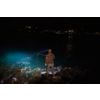 Personne debout sur des rochers au bord de l'eau, dos à la caméra, porte une chemise à carreaux et une casquette, tient un cadre métallique rectangulaire au-dessus de l'eau; une lumière bleue éclaire l'eau à gauche; lumières côtières lointaines à l'horizon