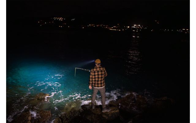 Personne debout sur des rochers au bord de l'eau, dos à la caméra, porte une chemise à carreaux et une casquette, tient un cadre métallique rectangulaire au-dessus de l'eau; une lumière bleue éclaire l'eau à gauche; lumières côtières lointaines à l'horizon
