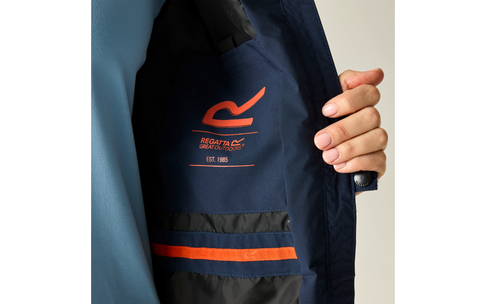 Innenansicht einer dunkelblauen Jacke; innen ein orangefarbenes Logo 'Regatta Great Outdoors' mit Text 'EST. 1985' und zwei horizontale Streifen (orange und dunkelblau) an der Innentasche; eine Hand hält die Jacke geöffnet