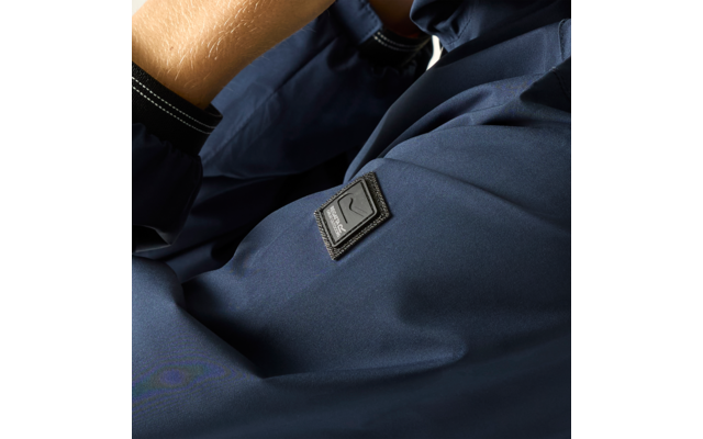 Nahaufnahme eines dunkelblauen Jackenärmels mit schwarzem quadratischem Logo-Patch, sichtbarem Unterarm und schwarzem Bündchen mit weißer Naht