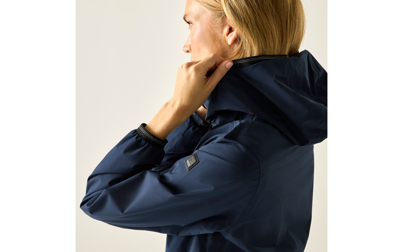 Seitliche Aufnahme einer Person mit blonden Haaren, die an der Kapuze einer dunkelblauen Jacke zieht; heller Hintergrund