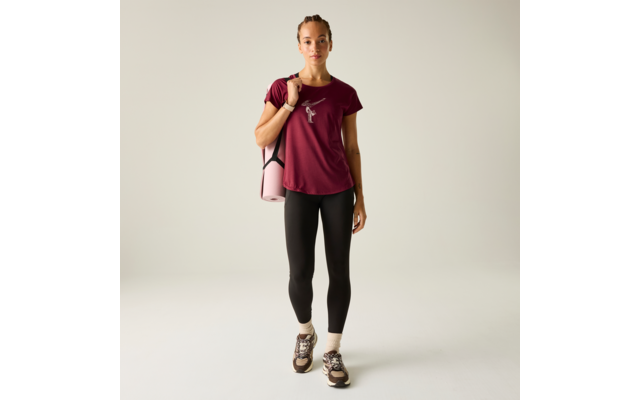 Person steht frontal, trägt bordeauxfarbenes T-Shirt mit kleinem weißem Logo, schwarze Leggings, Sportschuhe und hält eine zusammengerollte rosa Yogamatte über einer Schulter