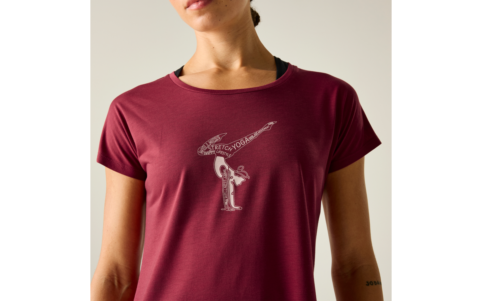 Oberkörper einer Person bis zum Kinn, trägt ein dunkelrotes T‑Shirt mit schwarzem Einsatz am Halsausschnitt. Auf der Brust ein kleines weißes Linienmotiv mit zwei stilisierten Figuren, eine nach vorn geneigt und eine waagrecht darüber