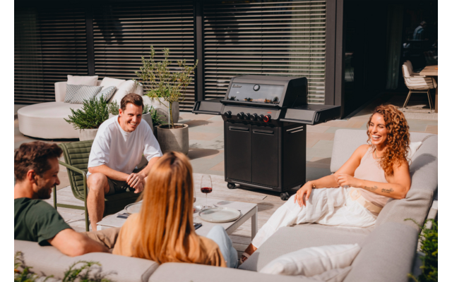 Vier Erwachsene sitzen auf Außensofas um einen niedrigen Tisch auf einer Terrasse; ein Gasgrill, Weingläser, Kissen und Pflanzen sind sichtbar