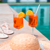 Zwei orangene Cocktails mit Eiswürfeln, Orangenscheiben und Strohhalmen auf dem Rand eines Schwimmbeckens, daneben eine Strohtasche und Sandalen