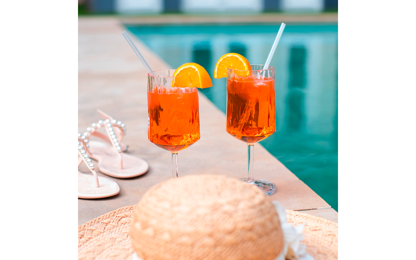 Zwei orangene Cocktails mit Eiswürfeln, Orangenscheiben und Strohhalmen auf dem Rand eines Schwimmbeckens, daneben eine Strohtasche und Sandalen