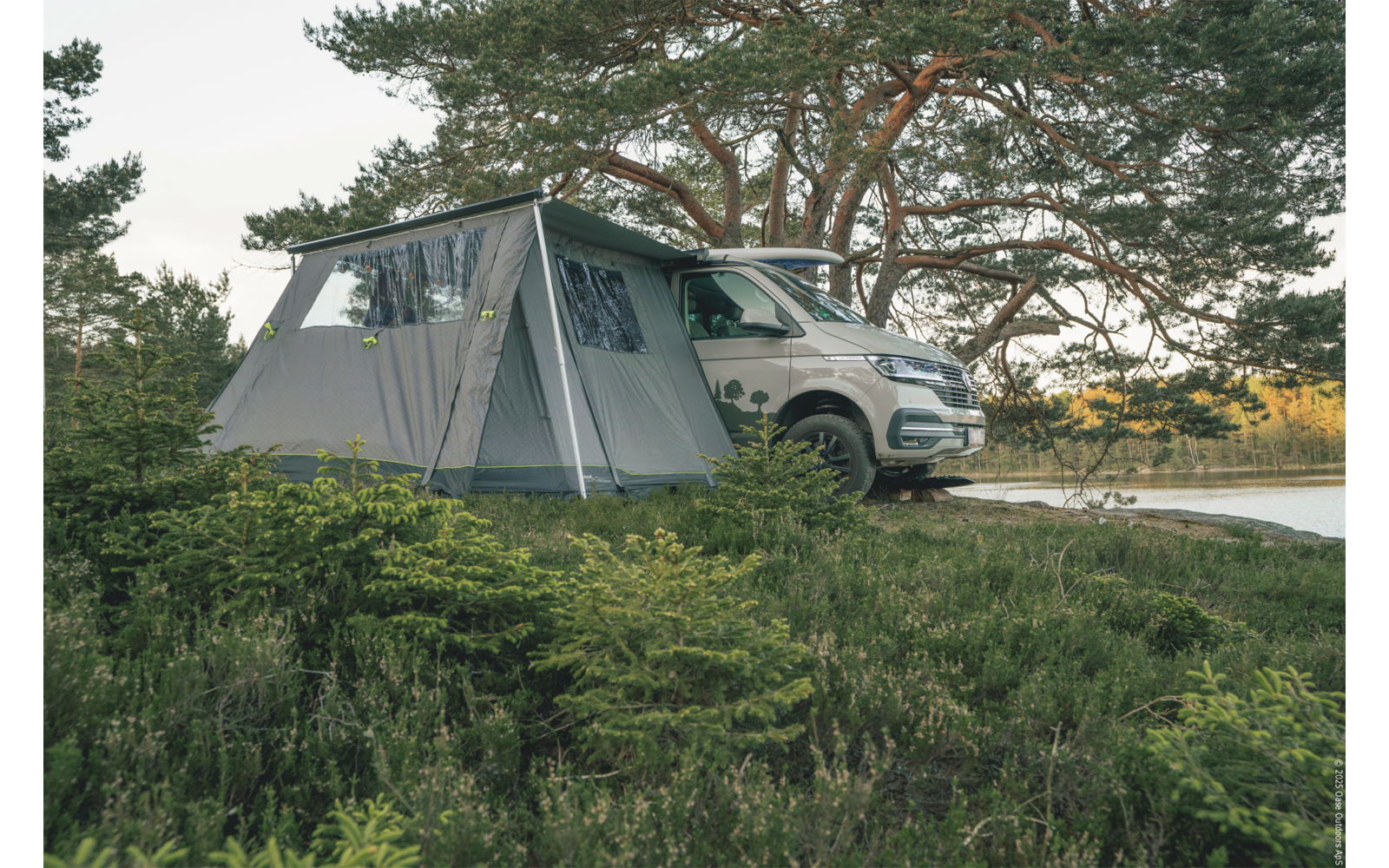 SUV bianco con una tenda grigia attaccata accanto a un lago, pini sullo sfondo, erba e arbusti bassi in primo piano