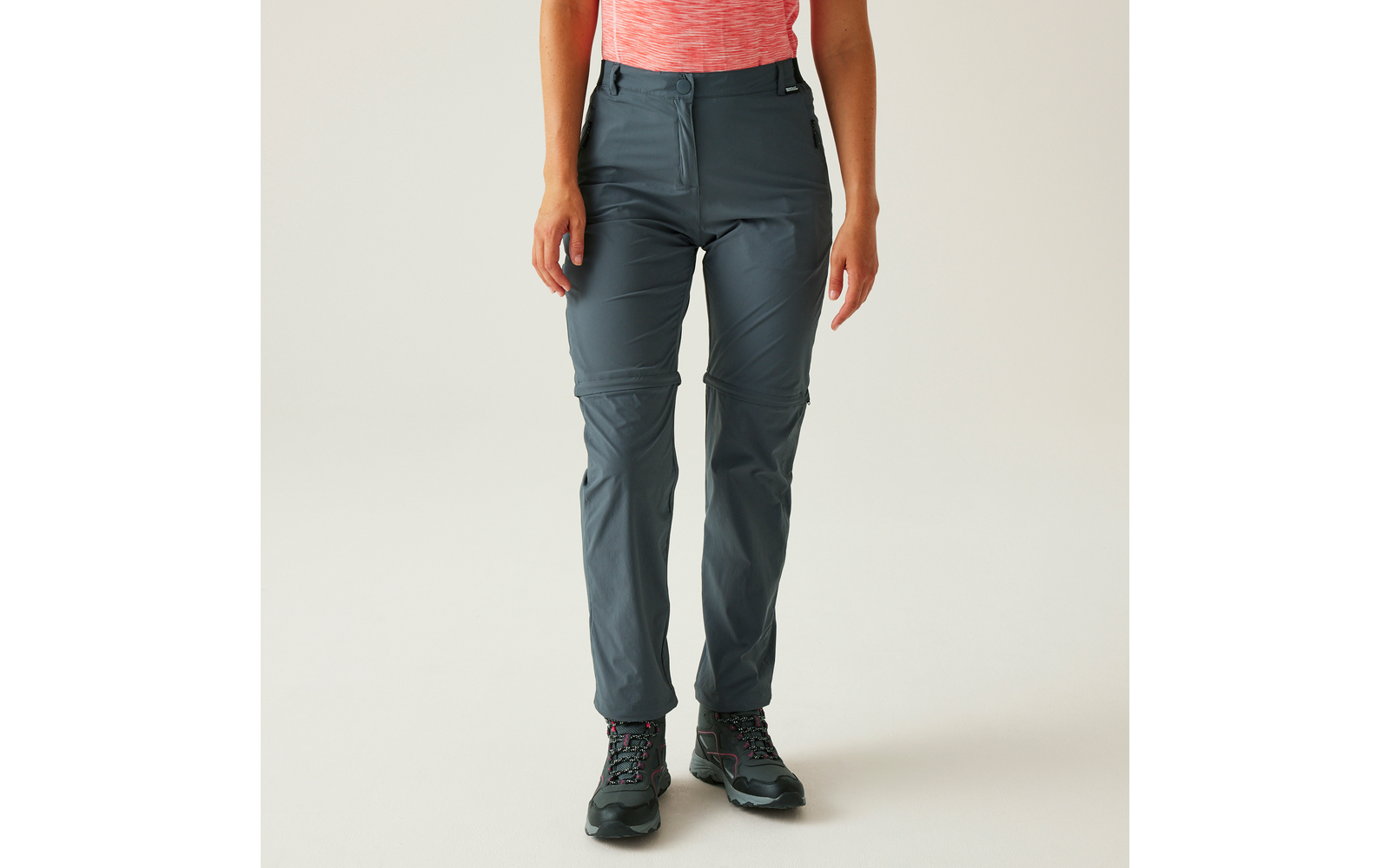 Unterkörper einer Person: graue lange Hose mit sichtbaren Reißverschlüssen an den Knien, Ansatz eines pink-orangefarbenen Oberteils und schwarze Schnürschuhe mit pinken Akzenten