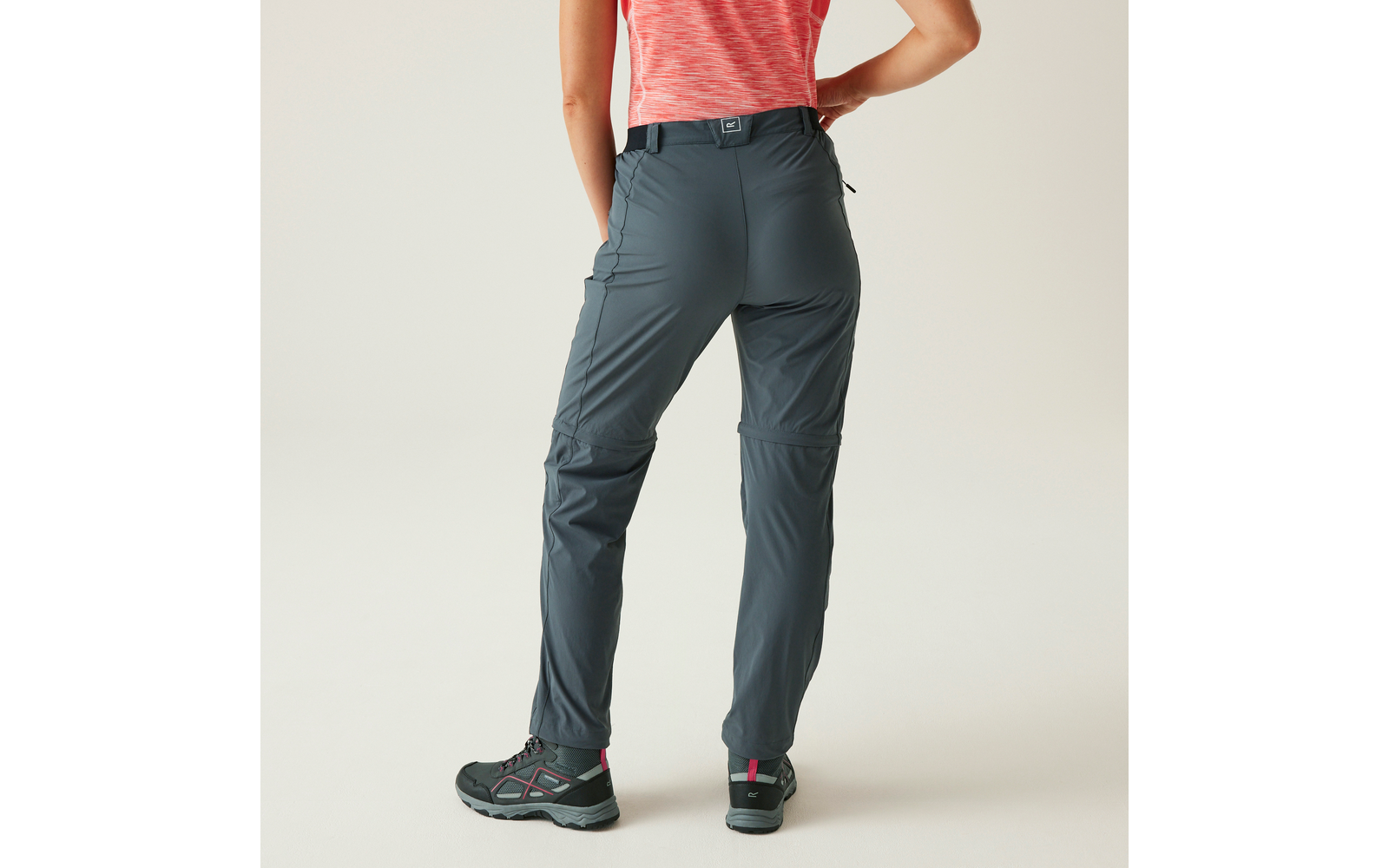 Rückenansicht einer Person, die dunkelgraue lange Hose mit Seitentaschen, korallenrotes T-Shirt und graue Schuhe mit pinken Details trägt; rechte Hand an der Hüfte; heller Hintergrund