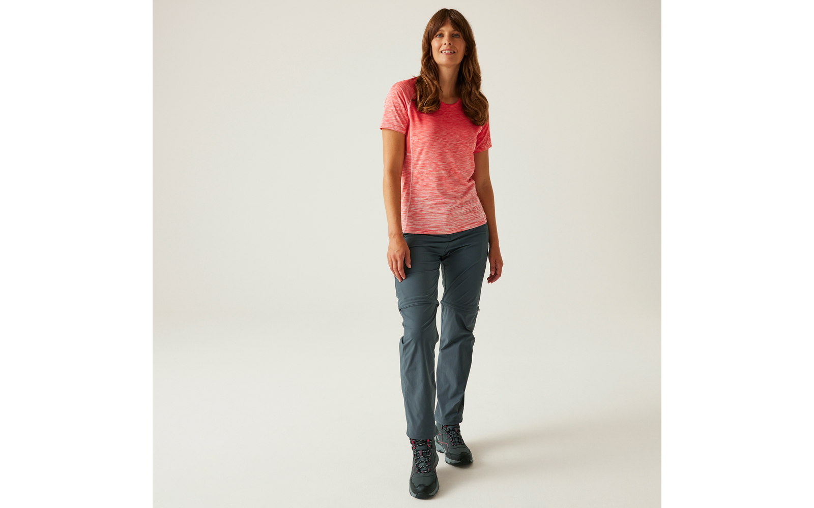 Frau mit langen braunen Haaren vor hellem Hintergrund, trägt rosa T‑Shirt, graue Wanderhose und dunkle Wanderschuhe