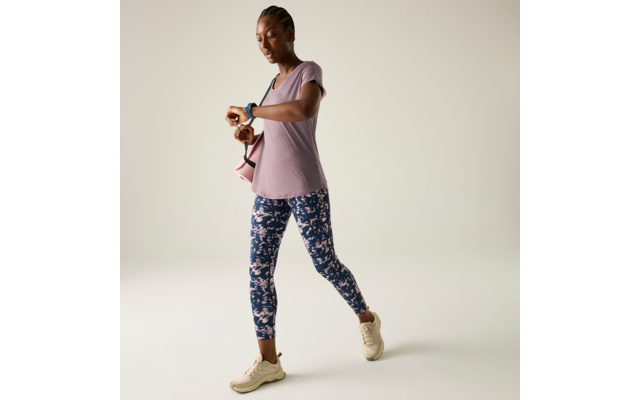 Eine Frau geht und schaut auf eine Smartwatch, trägt eine rosa Umhängetasche, ein hellviolettes T-Shirt, geblümte Leggings und cremefarbene Sneakers vor hellem Hintergrund