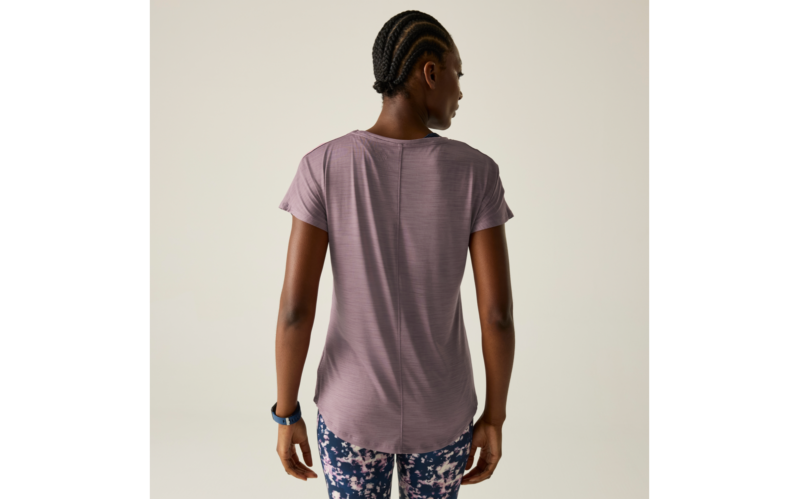 Person von hinten in violettem T-Shirt, gemusterten Leggings und Sportarmband vor hellem Hintergrund