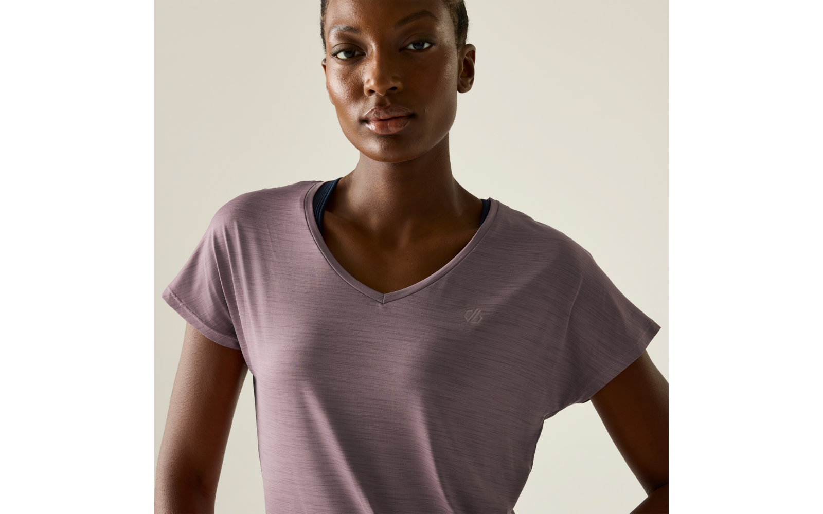 Person mit kurzem Haar, trägt ein fliederfarbenes V‑Ausschnitt‑T-Shirt mit kleinem Logo auf der Brust, sichtbare dunkle Träger darunter, neutraler Gesichtsausdruck, heller einfarbiger Hintergrund