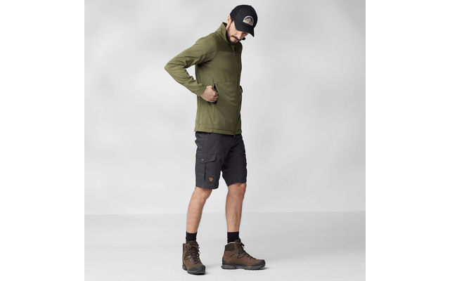 Mann in seitlicher Ganzkörperansicht, trägt olivgrüne Kapuzenjacke, dunkle Cargoshorts, braune Wanderstiefel und schwarze Kappe; eine Hand in Jackentasche; heller neutraler Hintergrund