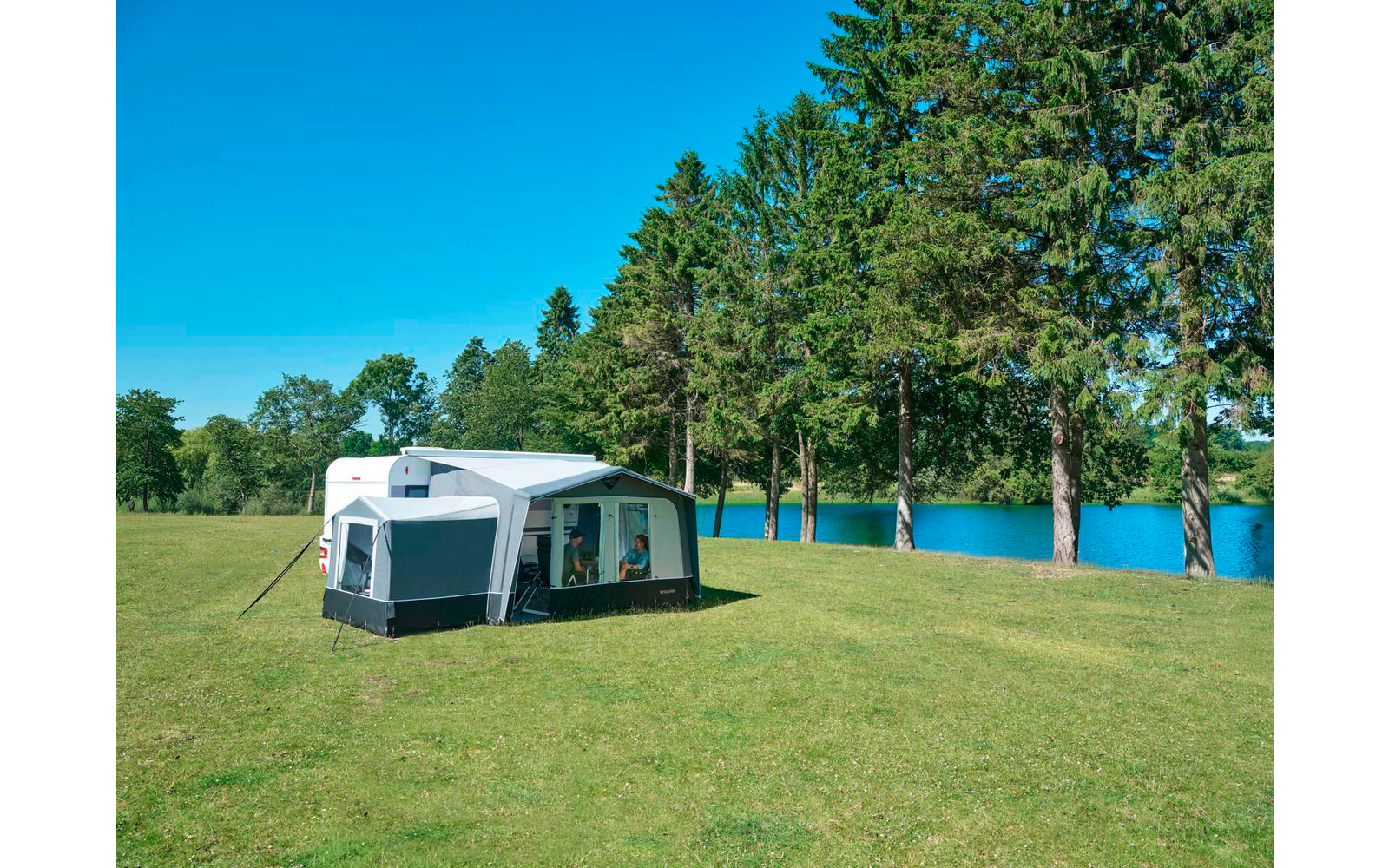 Weißer Wohnwagen mit angebautem grauem Vorzelt auf einer grünen Wiese, im Hintergrund hohe Kiefern und ein blauer See unter klarem Himmel