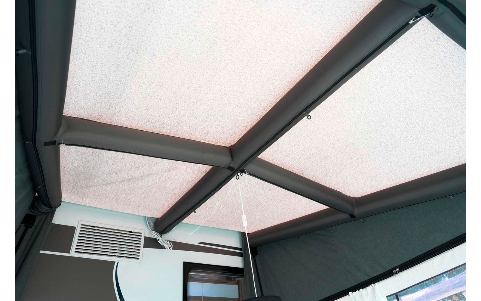Innenansicht eines hellen Stoffdachs mit schwarzen Metallträgern in Kreuzform, hängendem Zugband mit Gewicht, seitlicher Wand mit geschlitzter Lüftungsöffnung und Fenster mit Gardine