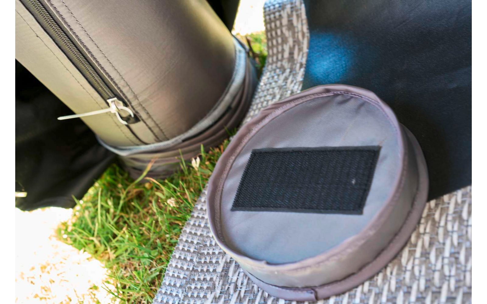 Runde graue Stofftasche mit schwarzem Klettfeld auf einer geflochtenen Matte, daneben ein schwarzer zylindrischer Gegenstand mit Reißverschluss und Gras