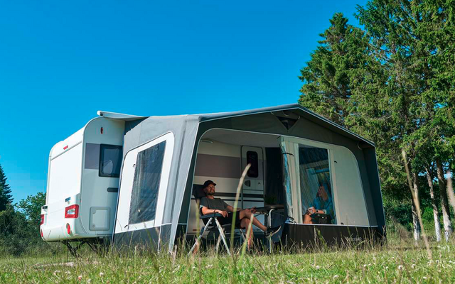 Weißer Wohnwagen mit angebautem Vorzelt auf grüner Wiese; zwei Personen sitzen an einem Tisch im Vorzelt; Bäume rechts; blauer Himmel