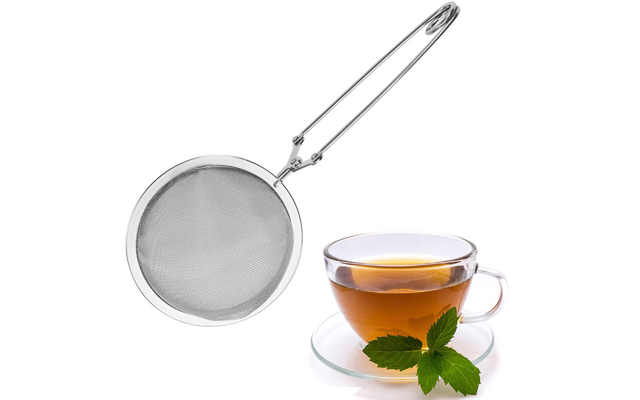Edelstahl-Teesieb mit langem Griff neben einer durchsichtigen Glas-Tasse mit Tee auf Untertasse, dazu frische Minzblätter