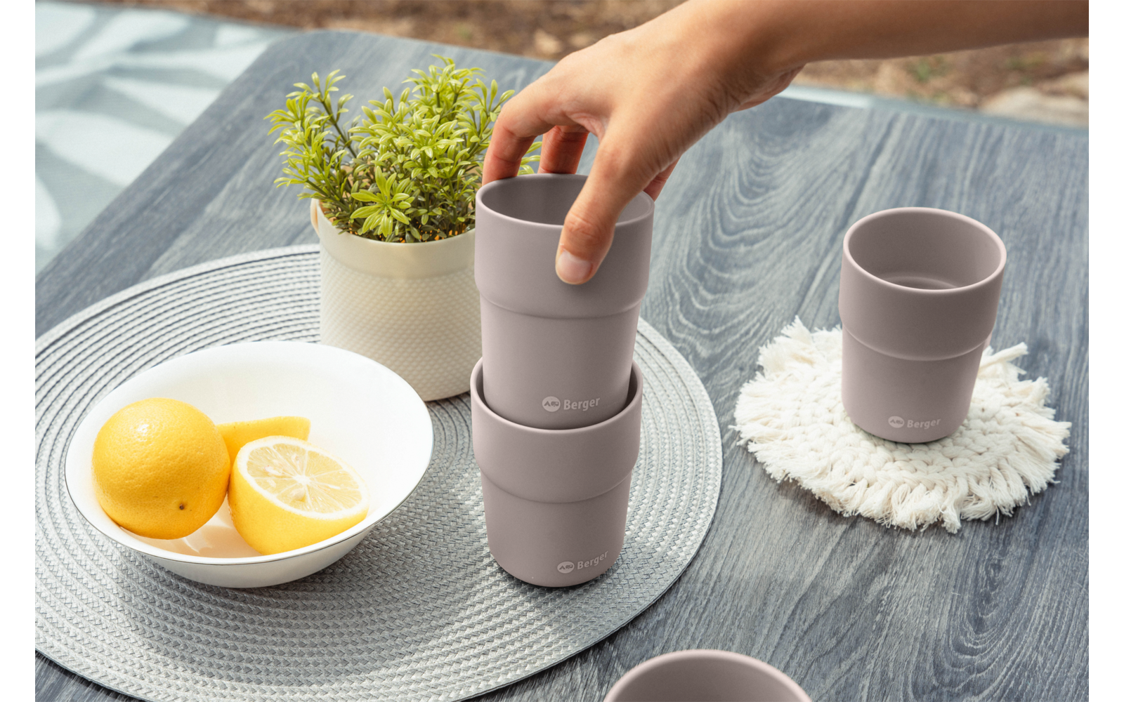 Hand hält einen grauen, stapelbaren Becher auf einem grauen Holztisch; daneben ein weißer Topf mit grüner Pflanze, eine weiße Schale mit ganzen und halbierten Zitronen und ein weiterer grauer Becher auf einer weißen Unterlage