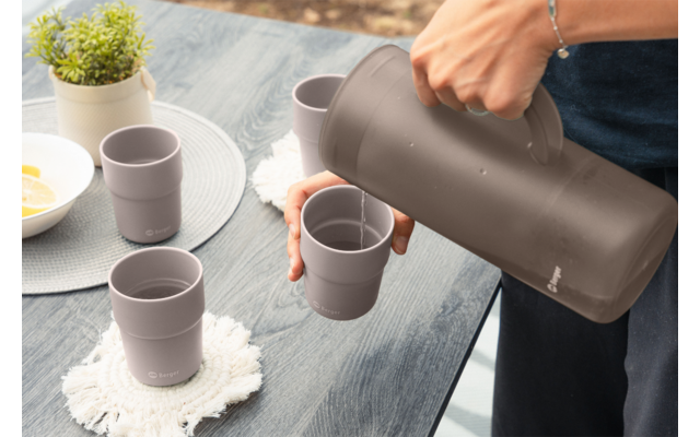 Hand gießt aus brauner Thermoskanne in braunen Becher auf dunklem Tisch; drei weitere Becher, zwei weiße Untersetzer, kleine Topfpflanze, weiße Schale mit gelben Scheiben