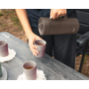 Hände einer Person in schwarzer Kleidung gießen aus einer dunkelbraunen Thermoskanne ein Getränk in einen rosafarbenen Becher auf einem grauen Holztisch; zwei weitere Becher mit Getränk auf dem Tisch