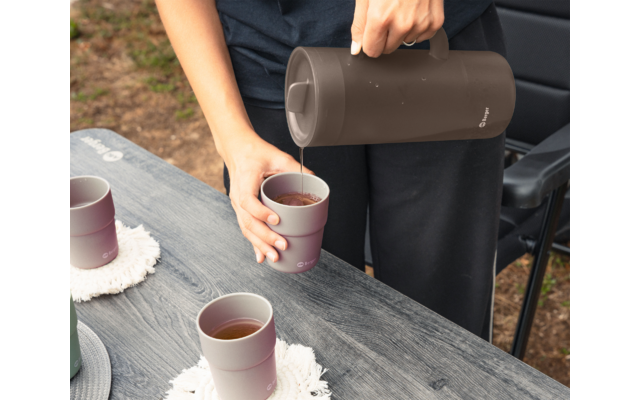 Hände einer Person in schwarzer Kleidung gießen aus einer dunkelbraunen Thermoskanne ein Getränk in einen rosafarbenen Becher auf einem grauen Holztisch; zwei weitere Becher mit Getränk auf dem Tisch
