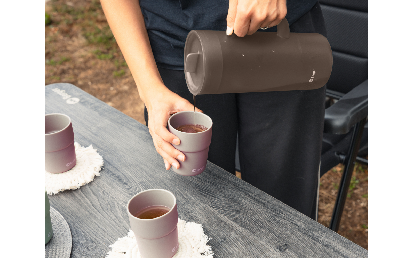 Hände einer Person in schwarzer Kleidung gießen aus einer dunkelbraunen Thermoskanne ein Getränk in einen rosafarbenen Becher auf einem grauen Holztisch; zwei weitere Becher mit Getränk auf dem Tisch