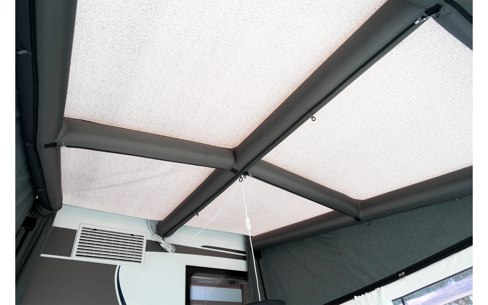 Decke mit hellen Stoffpaneelen, dunklen Querträgern, einer hängenden Schnur, einem rechteckigen Lüftungsgitter an der Wand und einem Fenster mit Rahmen