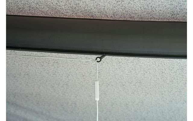 Grau gesprenkelte Decke mit einer dunklen horizontalen Leiste; an einer kleinen schwarzen Öse hängt eine weiße Schnur mit einem rechteckigen weißen Kunststoffgriff