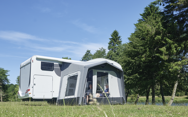 Caravana blanca con toldo gris sobre césped, árboles al fondo y cielo azul