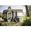 Frau steht neben offenem Campingbus mit ausgefahrener Markise, buntes Stand-up-Paddle-Board lehnt am Bus, See und Bäume im Hintergrund